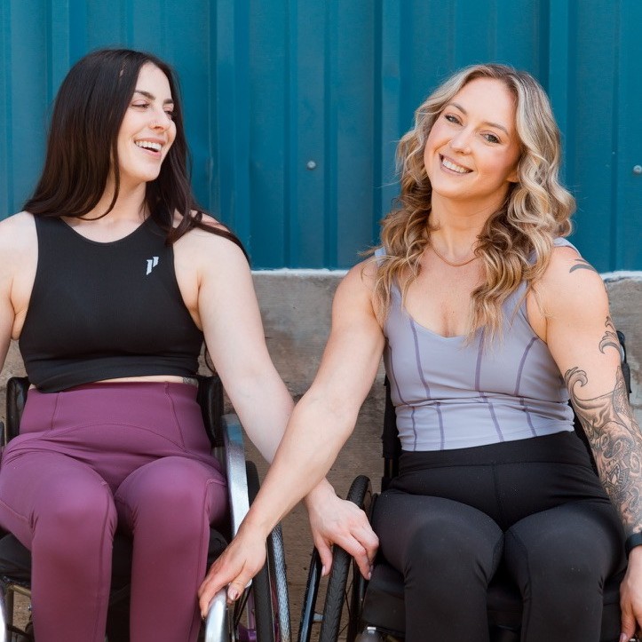 Nikki, with blonde hair smiles warmly at the camera wearing a black workout top, while Jesi, with brunette hair, smiles at Nikki wearing a blue workout top.