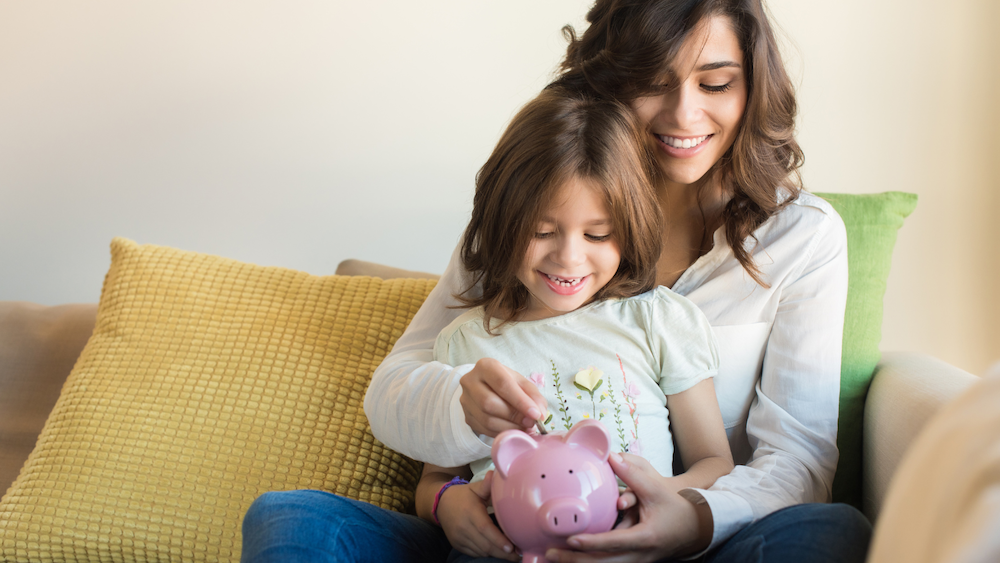 A woman sits on her couch, holding her daughter, as they drop a coin into a pink piggy bank.