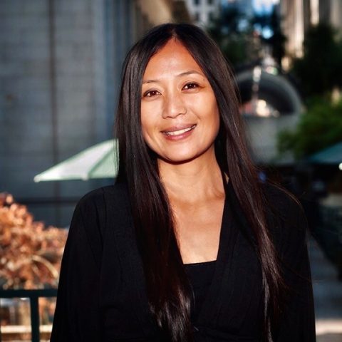 Reena De Asis smiles at the camera wearing a black camisole and black suit jacket.