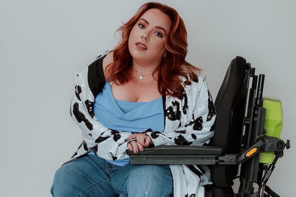 Marna poses for the camera in her wheelchair, wearing a blue top, jeans, black boots and a sequin cardigan.