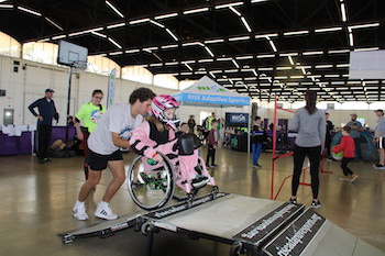 As young girl, clad in pink, is being pushed up a ramp for a wheelchair skate experience in the Rise Adaptive Sports Zone at Abilities Expo.