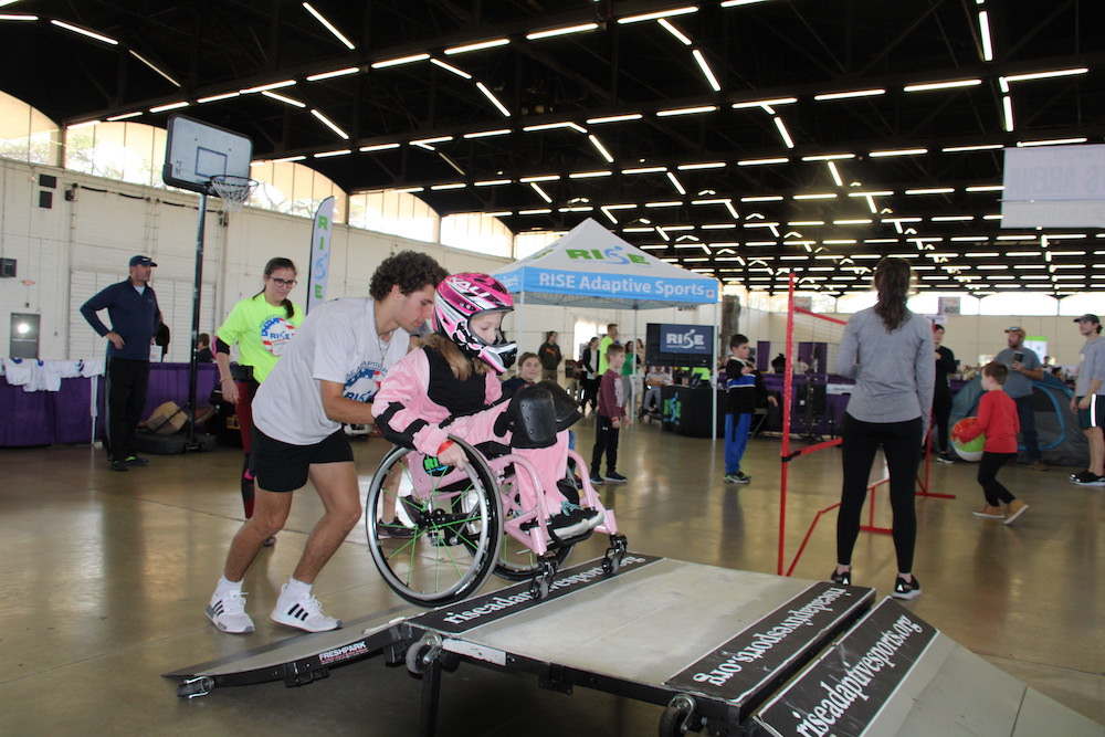 As young girl, clad in pink, is being pushed up a ramp for a wheelchair skate experience in the Rise Adaptive Sports Zone at Abilities Expo.