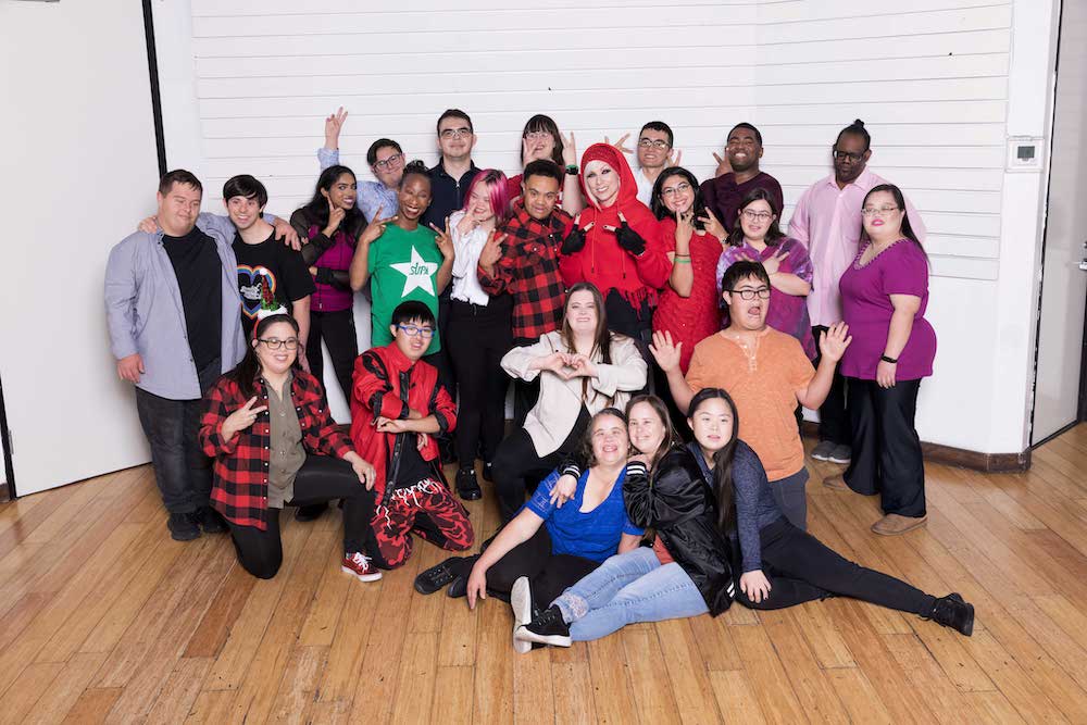 Founder of Straight Up Abilities, Robin Olive, smiles and makes a peace sign to the camera, surrounded by her group of talented dancers of all abilities and ages, all of which are smiling and laughing.