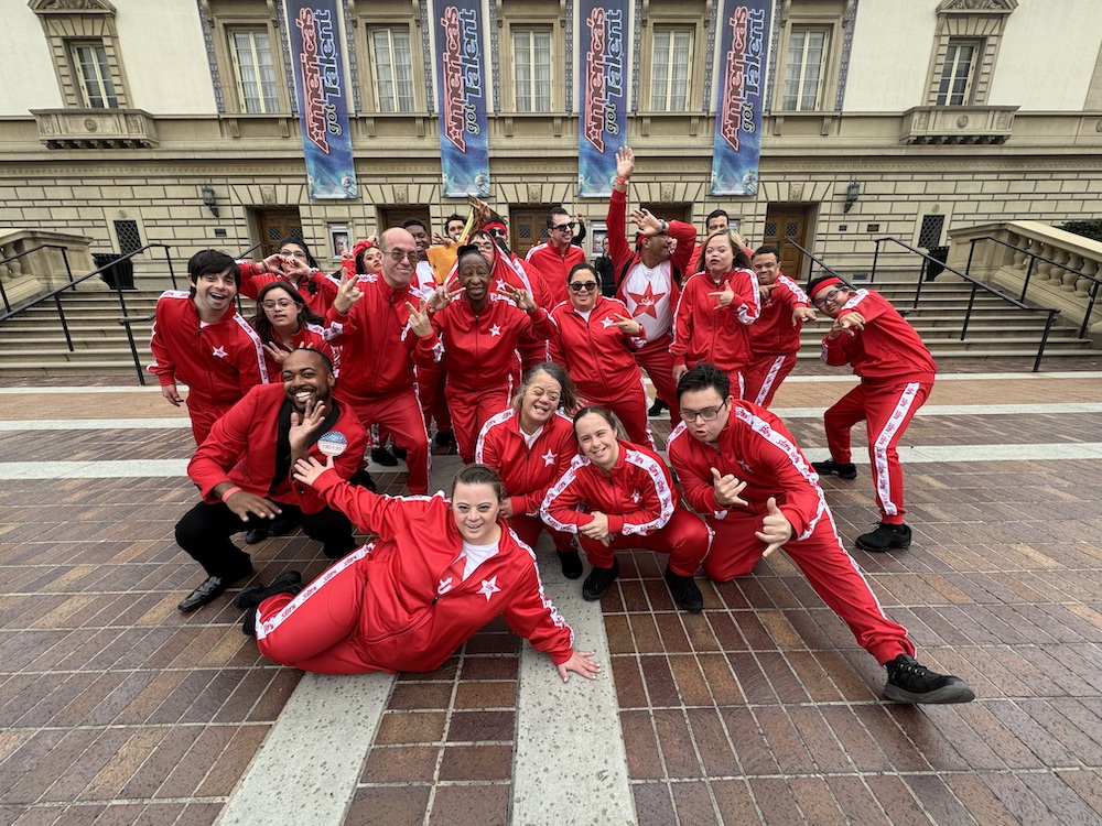 Founder of Straight Up Abilities, Robin Olive, smiles and makes a peace sign to the camera, surrounded by her group of talented dancers of all abilities and ages, all of which are smiling and laughing.