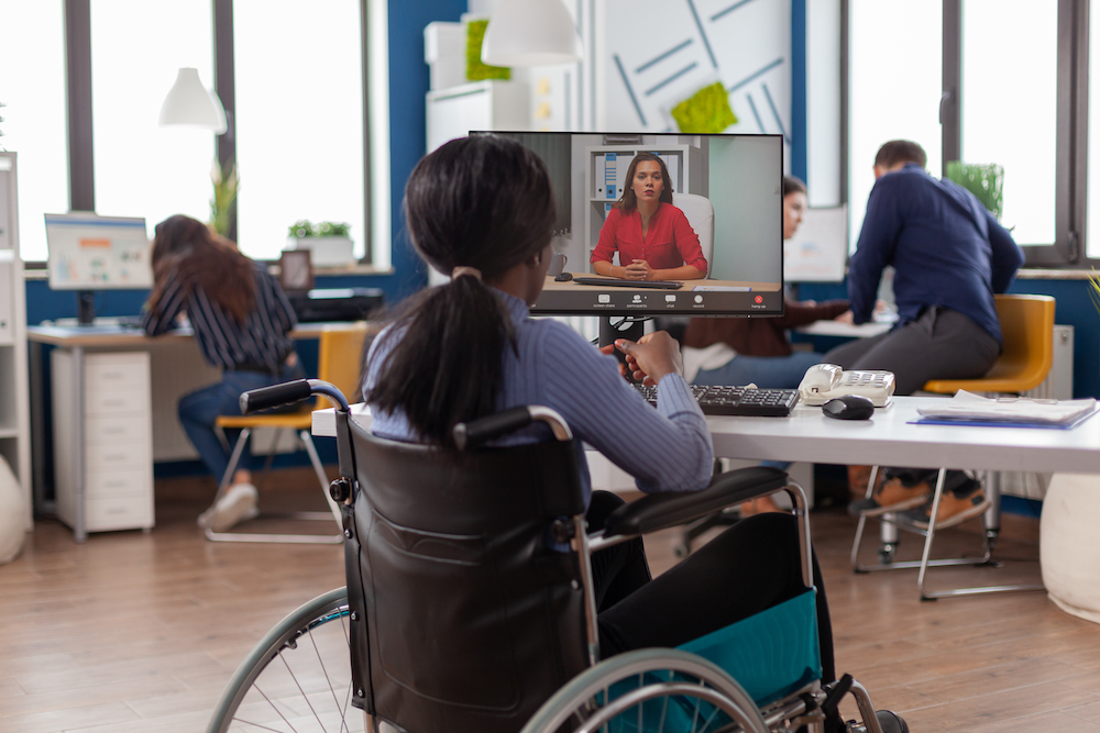 A disabled businesswoman is sitting in wheelchair talking with a remote partner on video call from startup business office.