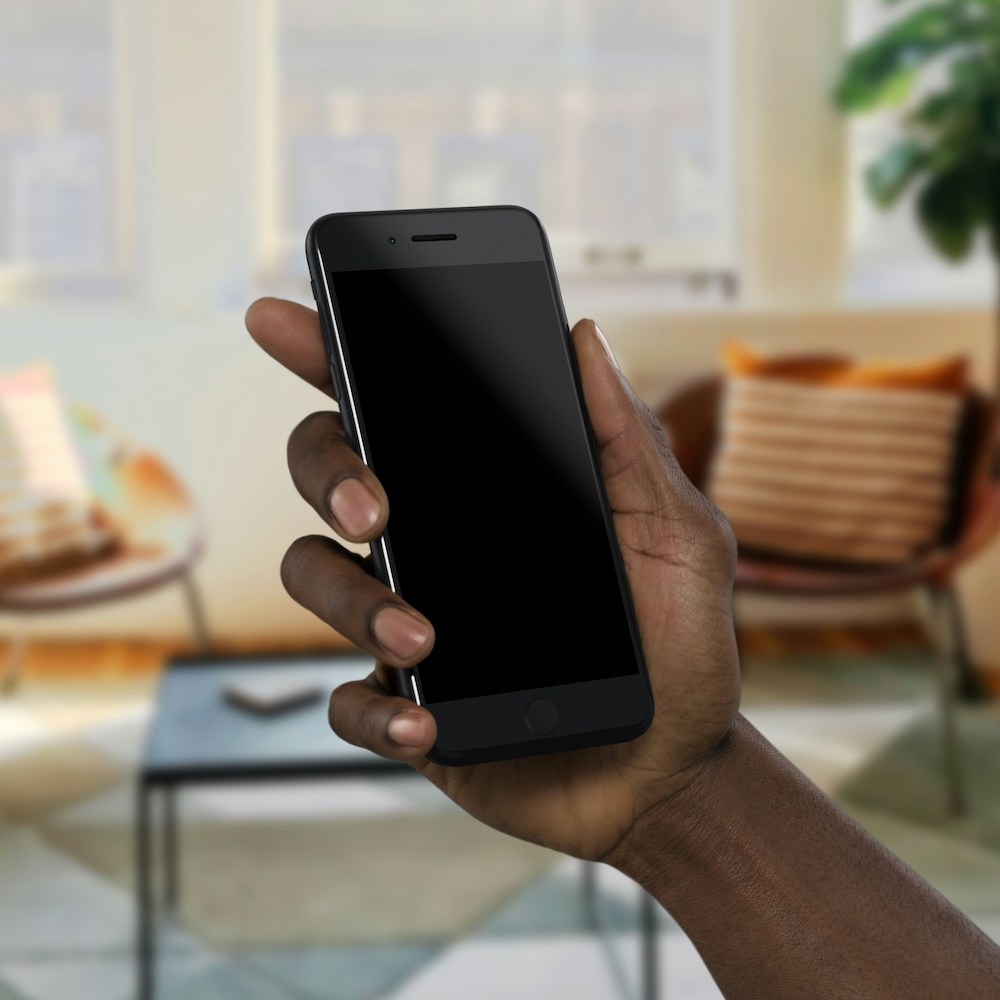 A hand is holding a cell phone with a living room in the background.