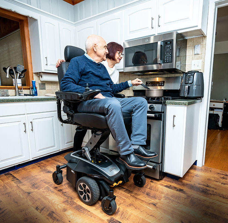 With his wide standing next to him, a man is cooking at a stove in the elevated position of his Pride Mobility Jazzy Air 2 Power Chair.