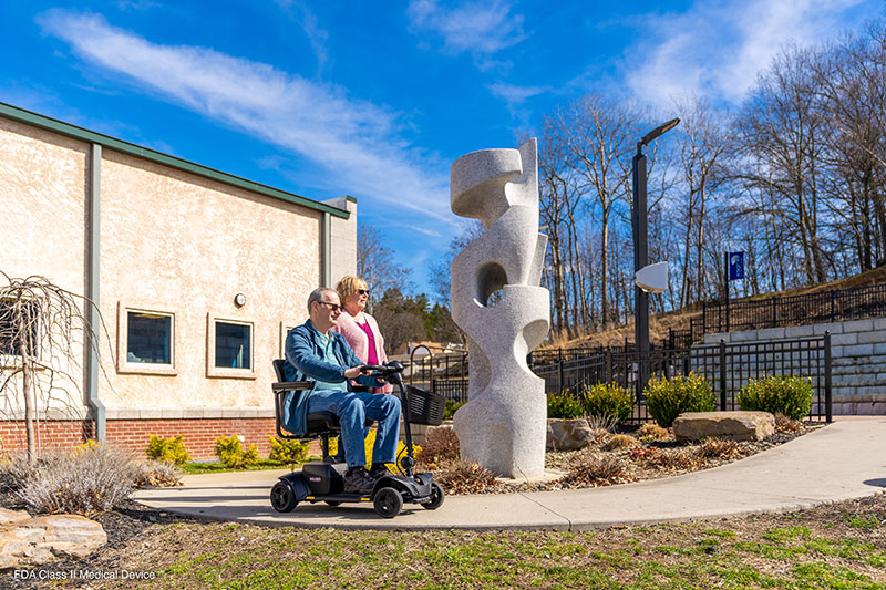 Sculpture: A man rides the Pride Mobility Go Go ES 2 electric scooter up the slight incline on a sidewalk as his wife walks alongside.