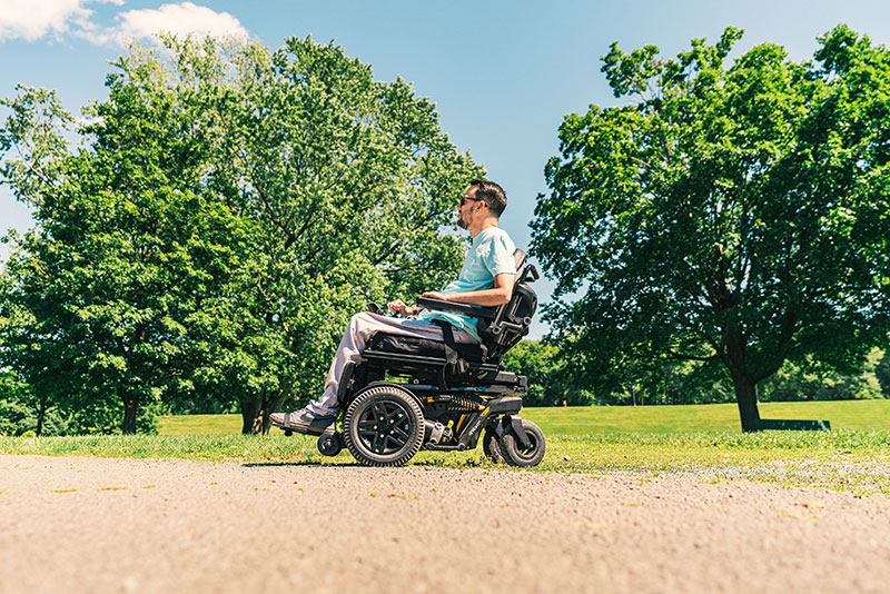 Profile view of a man riding a Quantum Rehab 4Front 2 power wheelchair along a dirt road with a green field and large trees in the background.