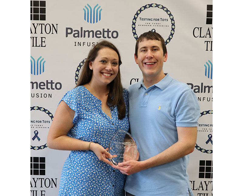 Dressed in blue, Brian and Tia Jones pose in front of a Palmetto Infusion step and repeat banner.