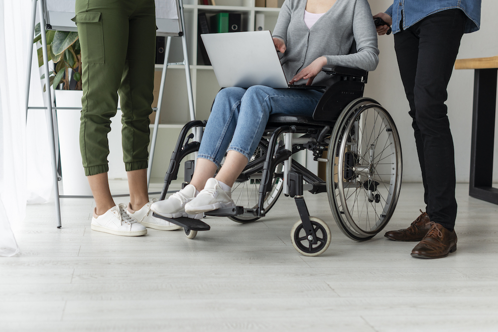 Three people are working together at the office with one using a wheelchair. No faces are visible.