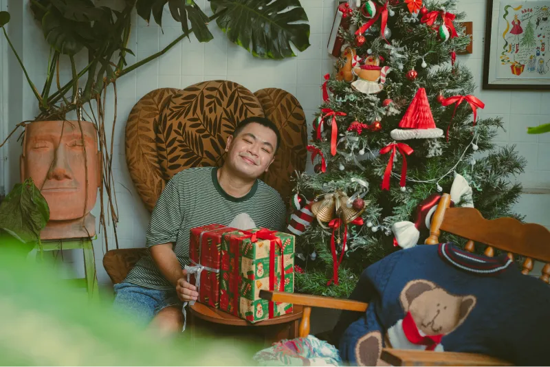 A young man with developmental disabilities is holding a holiday gift next to a festively decorated tree