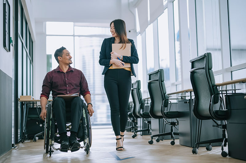 Two co-workers, one in a wheelchair and one nondisabled, chat as they walk/roll through the office.