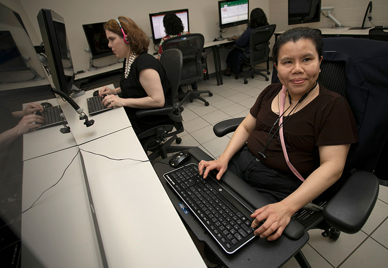 In an office with multiple computer stations, four women dressed in bussiness casual are sitting in office chairs in front of oversized monitors taking advantage of the assistive technology offered by East Texas Lighthouse for the Blind. Two women are also wearing headphones and one is pausing from her work and smiles in the direction of the camera.