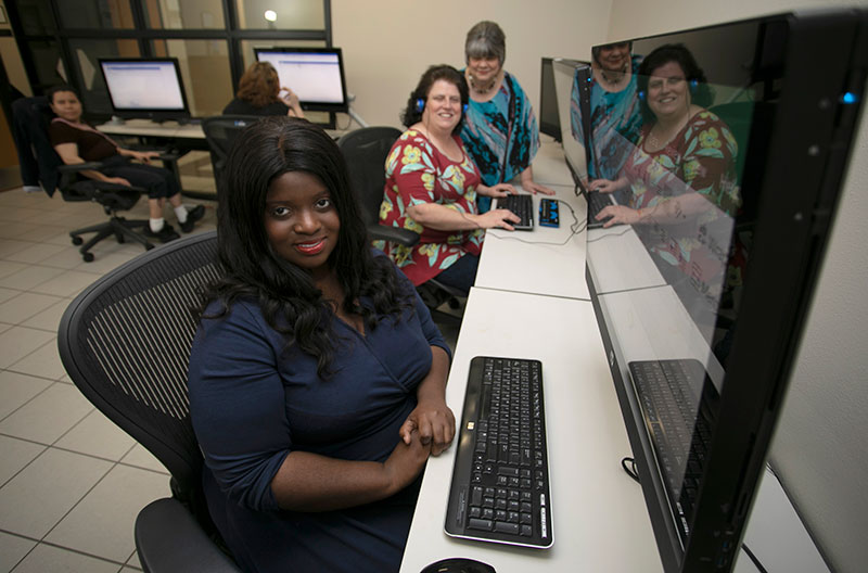 In an office with multiple computer stations and oversized monitors, four of the five women pictured pause from their work to smile in the direction of the camera. A braille keyboard sits in front of the keyboard of one of the stations.