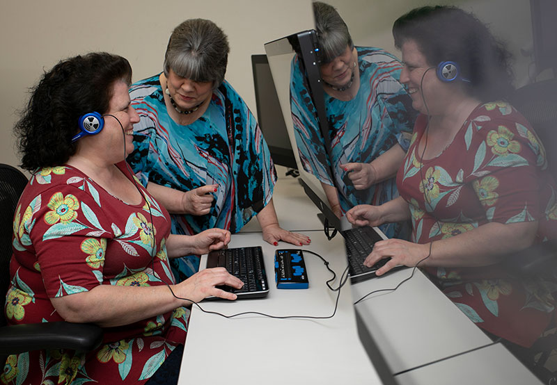 A woman dressed in a vibrant blue, red, gray and black shirt is standing next to a woman in a red dress with yellow and pale green flowers who is seated in front of computer keyboard, braille keyboard and oversized monitor. The seated woman is smiling as she received instruction from the standing woman.