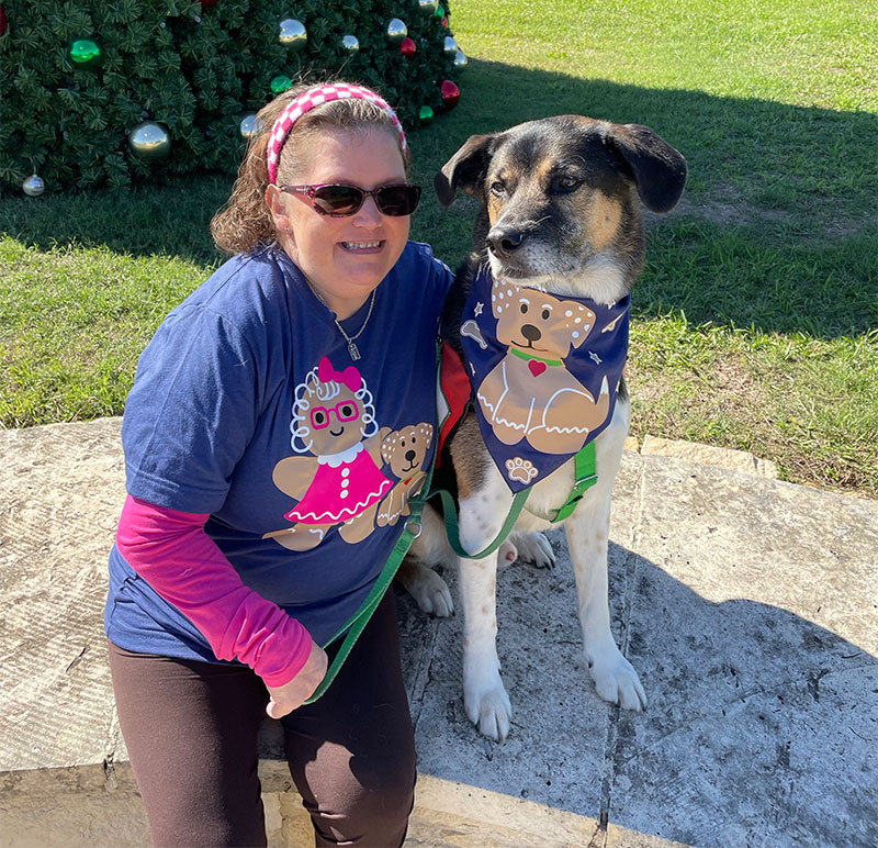 Amy Fey and Neo are sitting together outside on a sunny day. Her blue T-shirt has an illustration of her and Neo as a gingerbread person and dog. His bandana has a gingerbread dog.