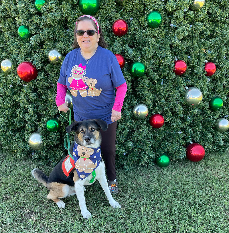 Neo is sitting in front of Amy as she stands in front of an outdoor Christmas tree. Her blue T-shirt has an illustration of her and Neo as a gingerbread person and dog. His bandana has a gingerbread dog.