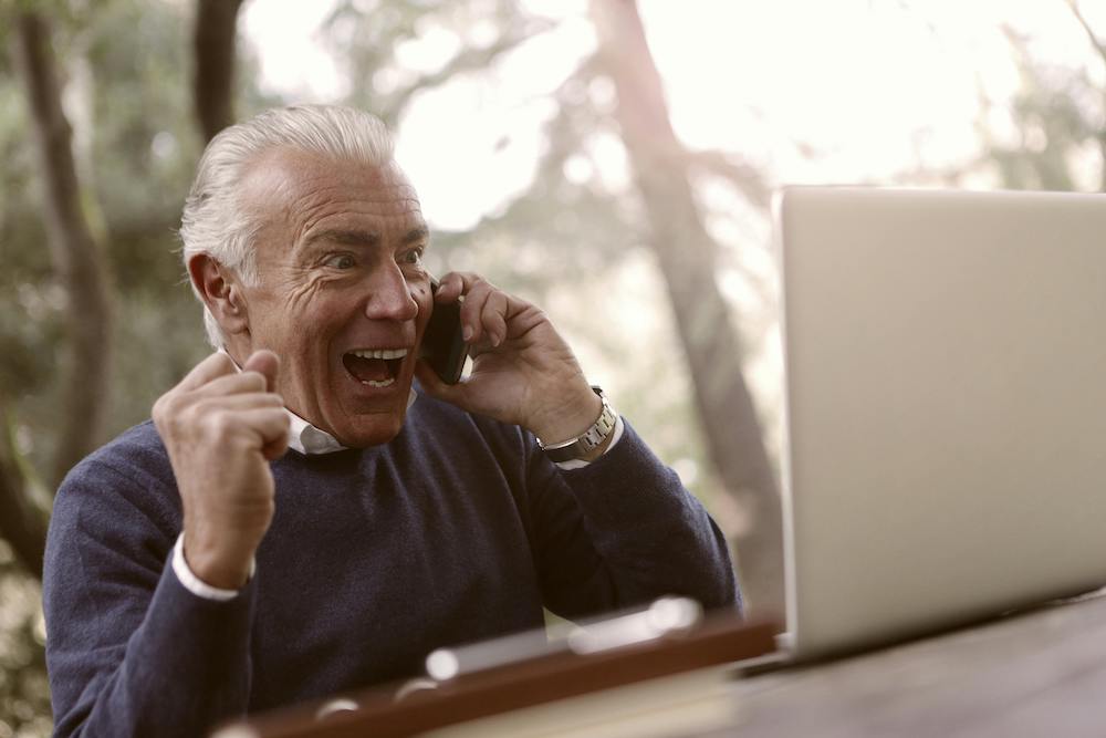 A senior is extremely excited by something on his laptop screen and talks on his cell phone.