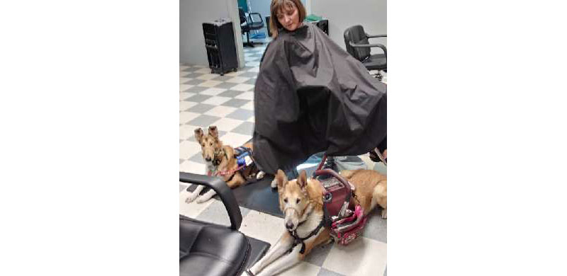 Clad in a black smock, Kristin Hartness sits in a chair at the salon as her two service dogs lay on the ground beside her.