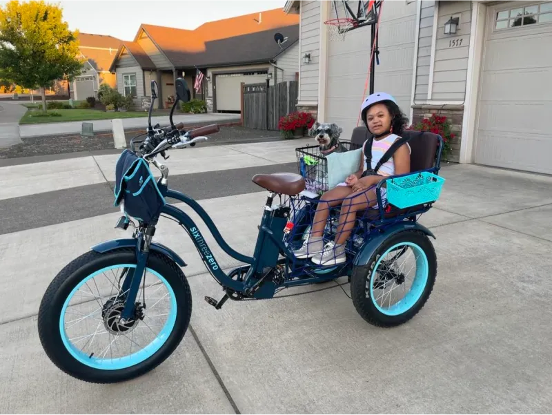 A young girl in a purple bike helmet is parked in the driveway waiting for her rider in the back of the sixthreezeo Electric Tricycle Rickshaw with a smiling passenger in the back.