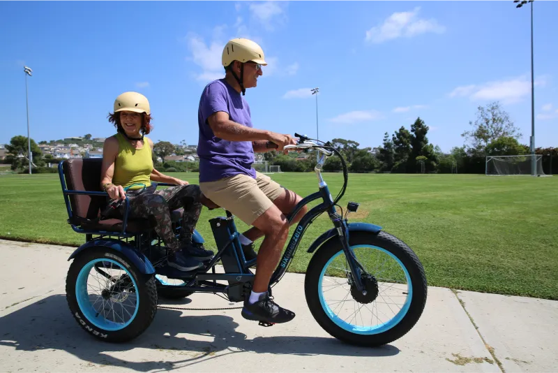 A man is riding his smiling wife past the soccer fields in a sixthreezeo Electric Tricycle Rickshaw.