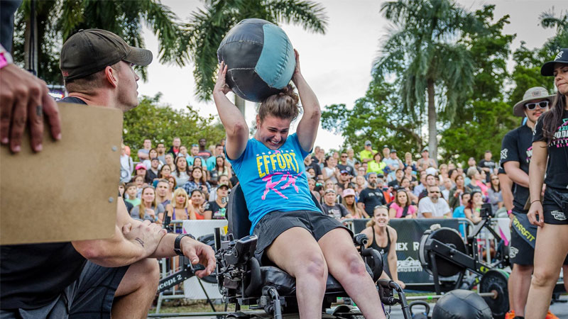 Female wheelchair user lifting heavy exercise ball above head.  