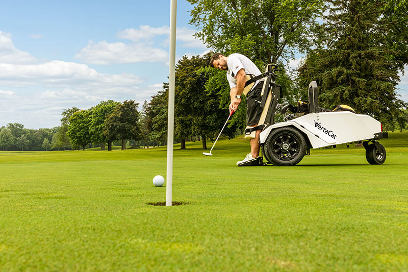 A golfer watches from a standing position with this VertaCat Stand-On-Command Mobility Rider as his putt rolls toward the hole. 