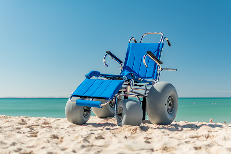 The bright blue Wheeleez Sandcruiser beach chair is by itself on a sandy beach on a sunny day with the blue-green ocean and blue cloudless sky in the background.