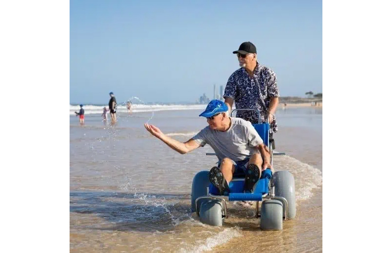 A senior man is pushing his senior friend on his Wheeleez Sandcruiser through the shallow water of the beach shoreline.