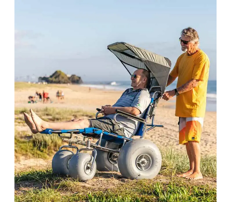 A senior is pushing his senior friend on his Wheeleez Sandcruiser through the grassy dune above the beach.