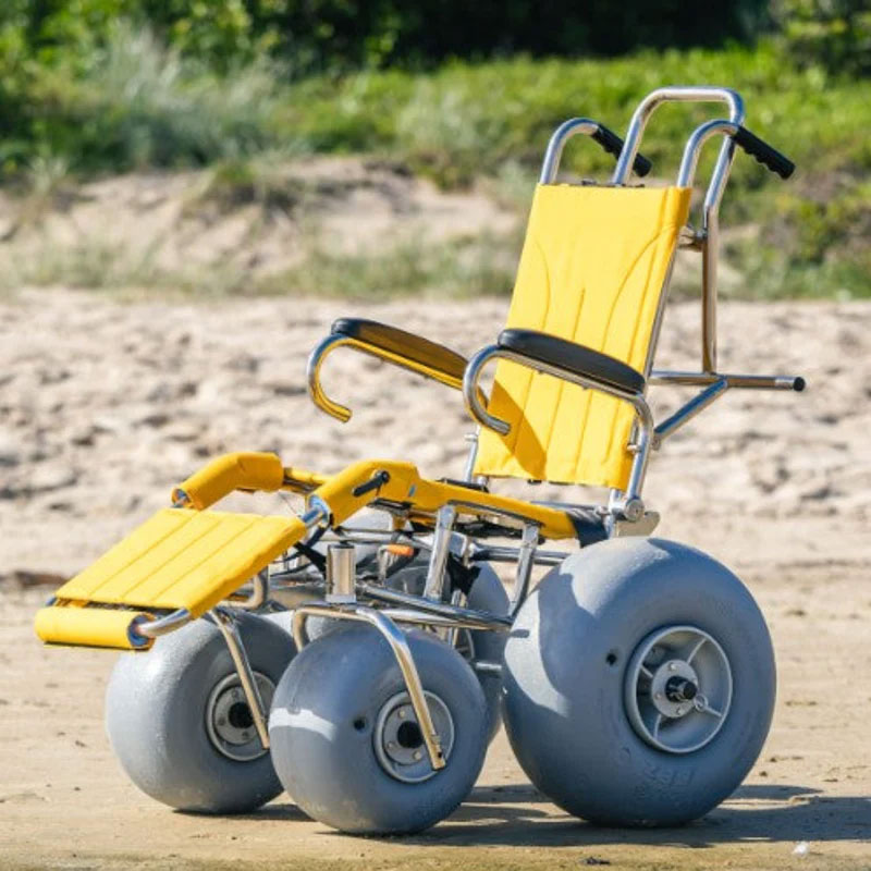 The bright yellow Wheeleez Sandpiper beach chair is by itself on a sandy beach with grassy dunes in the background.