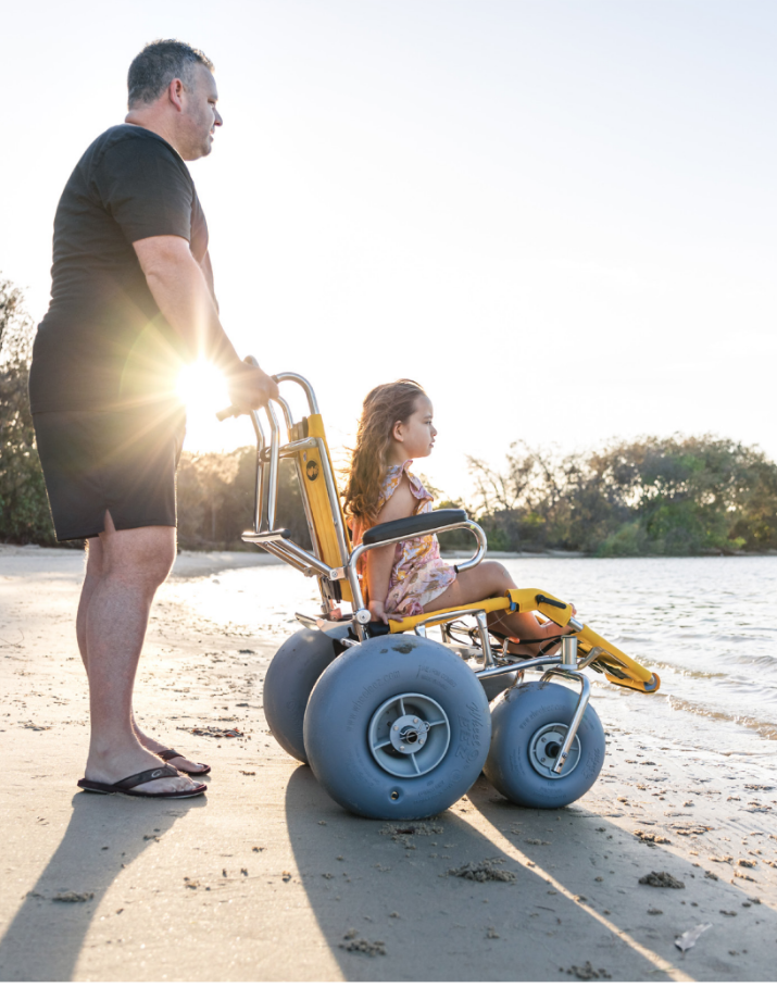 A young girl in a Wheeleez beach wheelchair and her father standing behind her are booth looking out over the water.