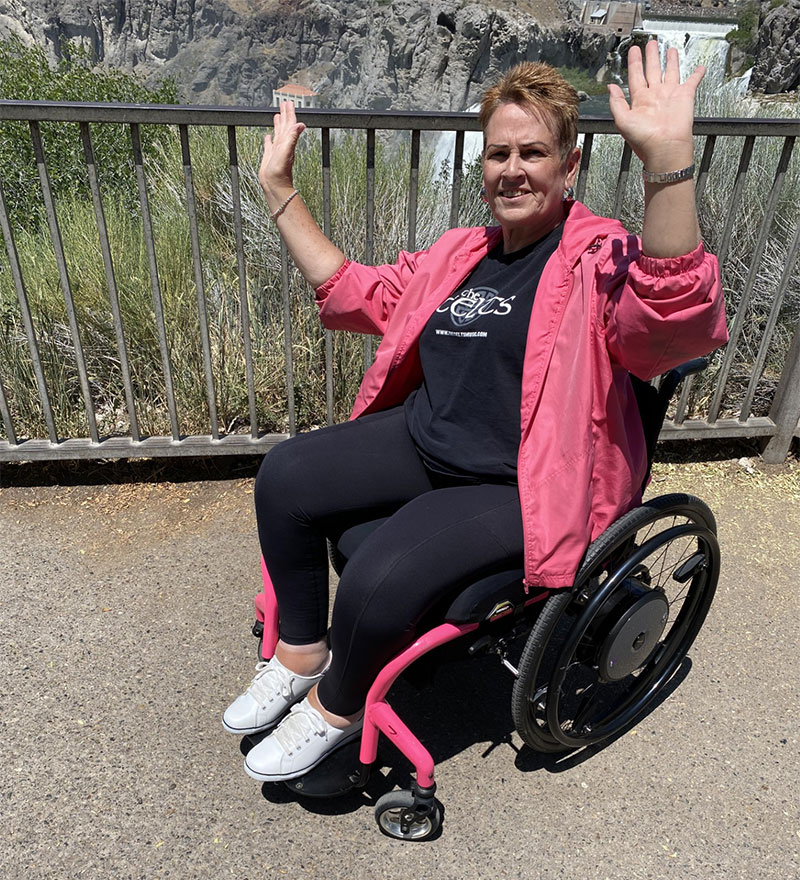 A smiling woman in a pink jacket and wheelchair with raised hands is sporting her Zip Hers tights.